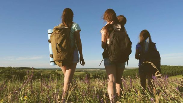 Happy mom and daughters, a little baby travel across the field with backpacks in colors in the summer. Family life. Tourist girls and a little baby go camping in the forest. Teamwork travelers - Photo, Image