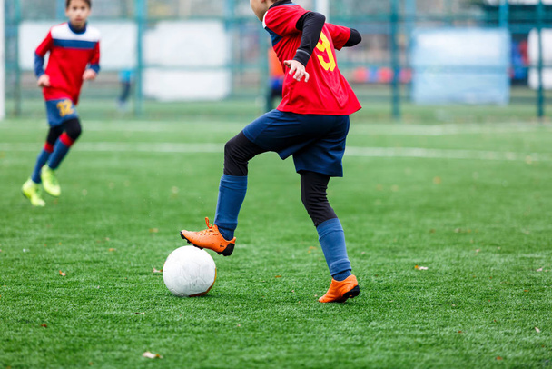 赤と青のスポーツウェアの少年たちは、フィールドにサッカー、ドリブルボールを果たしている。緑の芝生の上でボールを持つ若いサッカー選手。トレーニング、サッカー、子供のためのアクティブなライフスタイルの概念 - 写真・画像