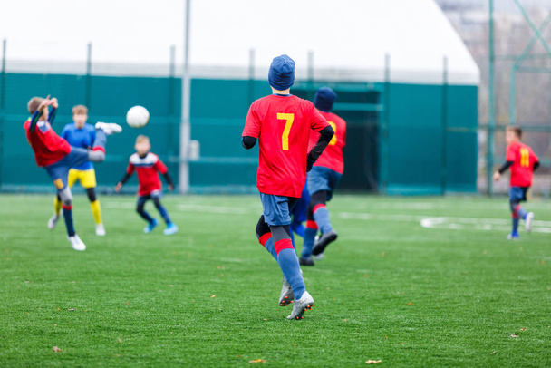 Chłopcy w czerwonym i niebieskim stroju sportowym grają w piłkę na boisku, dryblują. Młodzi piłkarze z piłką na zielonej trawie. Trening, piłka nożna, aktywny tryb życia dla dzieci - Zdjęcie, obraz
