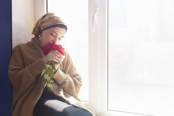 Jeune femme musulmane assise sur le rebord de la fenêtre avec bouquet de fleurs dans les mains. Femme du Moyen-Orient déprimée près de la fenêtre à la maison. Seule fille sans pom girl se sentant stressée et attendant
 - Photo, image