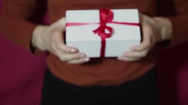 Beautiful girl holds out a gift in a white box ,with a red ribbon. Beautiful girl hands hold a New Years gift, in a white box with a red ribbon. Gift for the holiday. - Filmagem, Vídeo