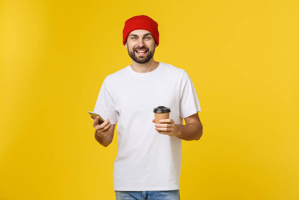 Arckép vidám fiatal férfi alkalmi ruhában, állandó izolált át a sárga háttér gazdaság mobiltelefon, elvihető kávéfogyasztás. - Fotó, kép