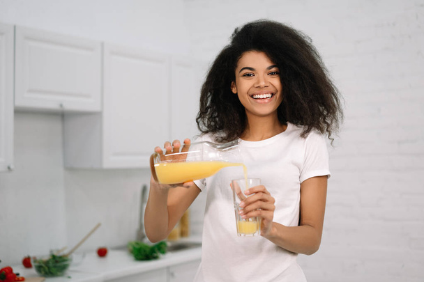 Νεαρή όμορφη Αφρο-Αμερικανίδα που κρατάει το γυαλί με έναν χυμό πορτοκαλιού, κοιτάζει την κάμερα και χαμογελάει. Υγιεινός τρόπος ζωής έννοια, φυσικό βιολογικό πρωινό στην κουζίνα  - Φωτογραφία, εικόνα