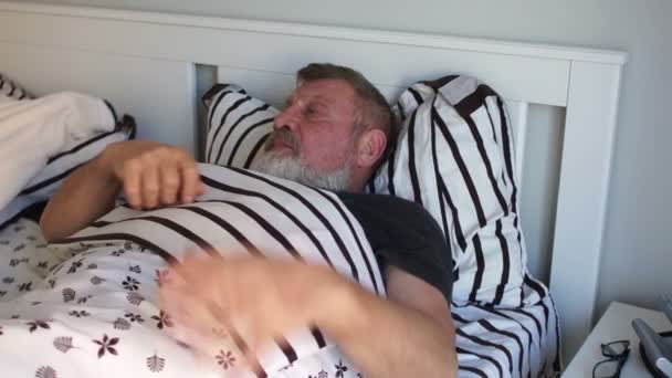 An elderly man with a beard wakes up and gets out of bed in the morning, stretches, holds on to his sore shoulder - 映像、動画