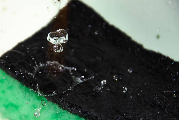 Zelená houba na mytí nádobí leží na bílém pozadí a proud vody na ni teče velmi blízko, makro. Spray visí ve vzduchu. Makro vody a houby - Fotografie, Obrázek