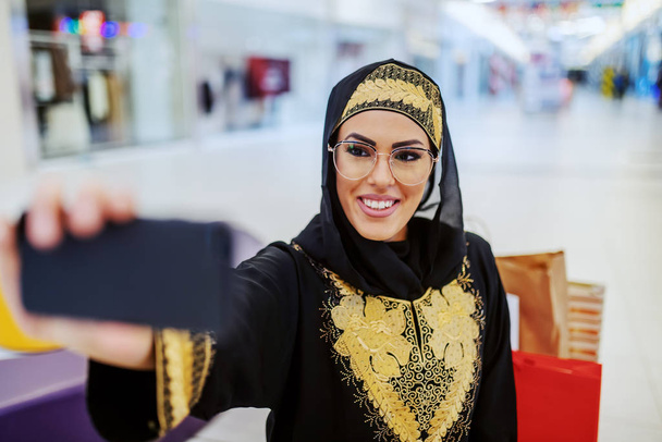 Ohromující mladá arabka v tradičním oblečení s krásným zubatým úsměvem stojící v nákupním centru a brát selfie pro sociální média. Generace tisíciletí. - Fotografie, Obrázek