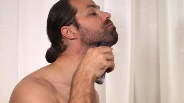 Un joven guapo con el pelo largo se afeita la barba con una podadora en el baño. Primer plano
. - Metraje, vídeo