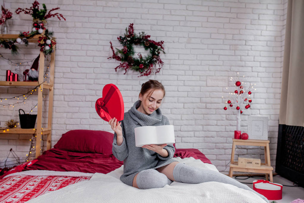 Portrait of a girl in a grey sweater with a heart-shaped red gift box sitting in the apartment, Valentine's day concept, copy space - Photo, image