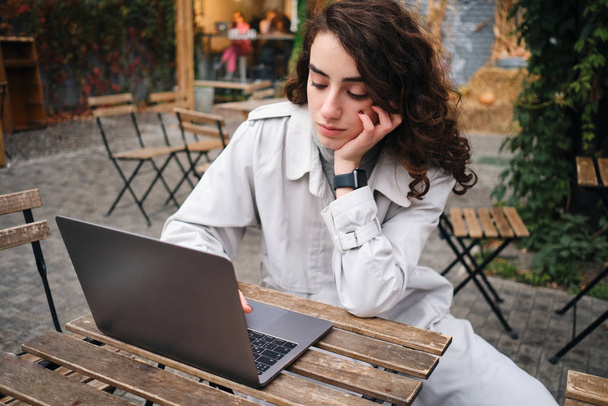 Hermosa chica morena casual en gabardina cuidadosamente trabajando en el ordenador portátil en la cafetería al aire libre
 - Foto, imagen