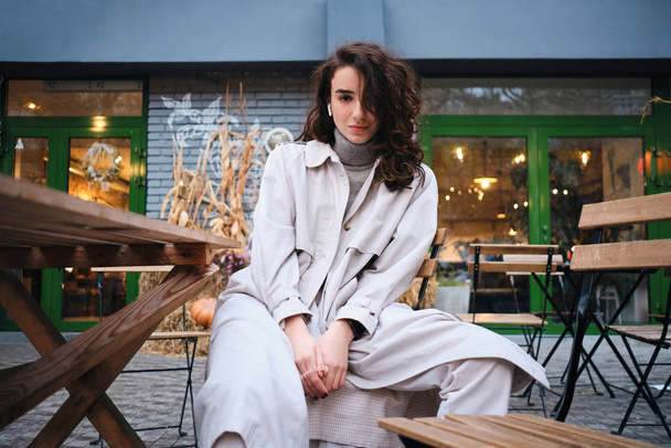 Menina morena elegante bonita em casaco de trincheira sonhadoramente olhando na câmera no café na rua
 - Foto, Imagem