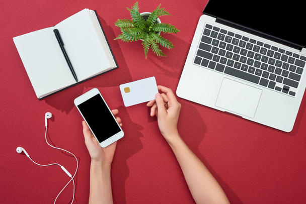 cropped view of woman holding credit card and smartphone near laptop, earphones, pen, notebook and plant on red background - Fotó, kép