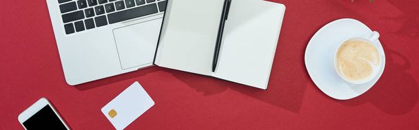 κορυφαία άποψη της πιστωτικής κάρτας σε κόκκινο φόντο με smartphone, laptop, καφέ, notebook με στυλό, πανοραμική λήψη - Φωτογραφία, εικόνα