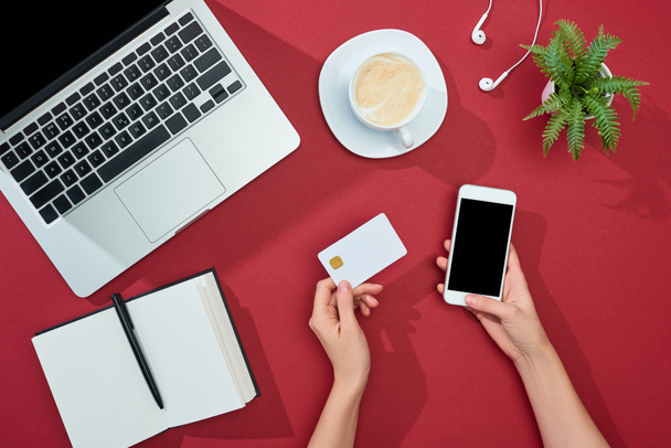 vista parcial de la mujer que sostiene la tarjeta de crédito y el teléfono inteligente cerca de café, ordenador portátil, auriculares, portátil y planta sobre fondo rojo
 - Foto, Imagen