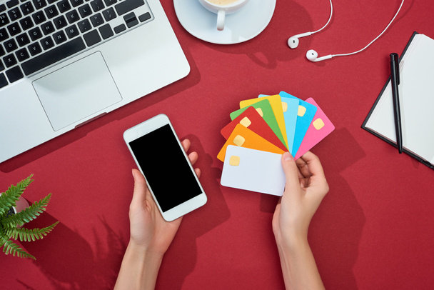 oříznutý pohled ženy držící pestrobarevné prázdné kreditní karty a smartphone na červeném pozadí s notebookem, sluchátky a kávou - Fotografie, Obrázek
