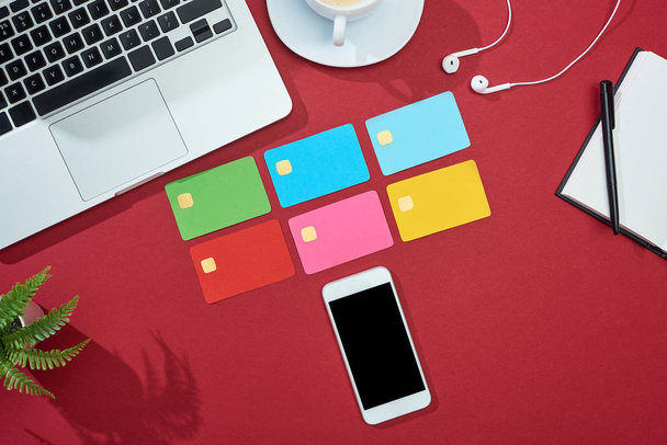 top view of multicolored empty credit cards on red background with smartphone, laptop, earphones and coffee - Foto, imagen