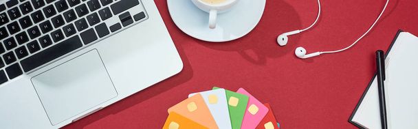 top view of multicolored empty credit cards on red background with laptop, earphones and coffee, panoramic shot - 写真・画像