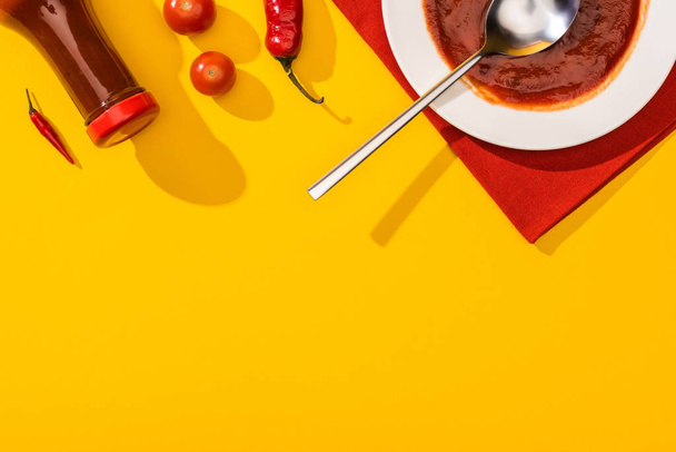 Top view of chili peppers with cherry tomatoes, plate and bottle with ketchup on yellow surface - Φωτογραφία, εικόνα