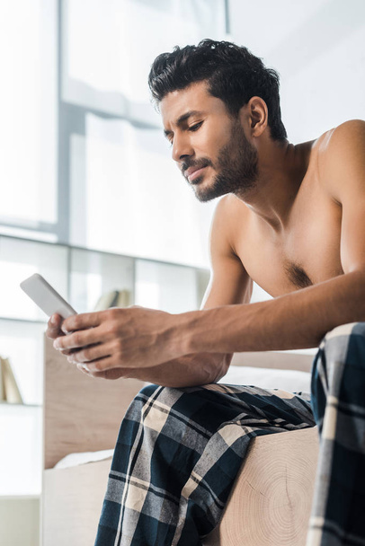 handsome and sexy bi-racial man sitting on bed and using smartphone in morning  - Photo, Image