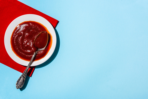 Horní pohled na chutnou rajčatovou omáčku v talíři s lžící na červeném ubrousku na modrém povrchu - Fotografie, Obrázek