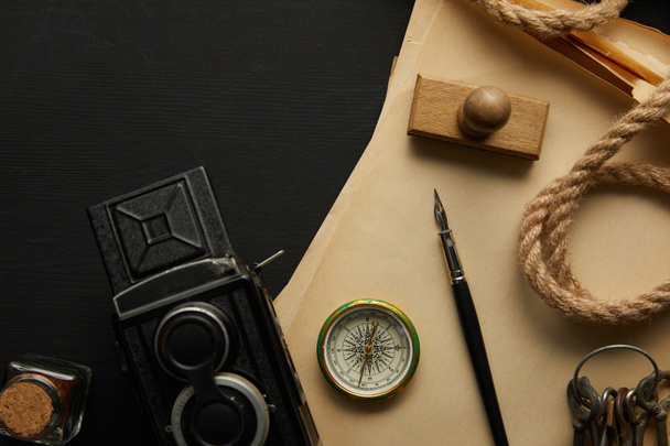 κορυφαία άποψη του vintage κάμερα, χαρτί, πένα στυλό, πυξίδα, σφραγίδα και τα κλειδιά σε μαύρο φόντο - Φωτογραφία, εικόνα