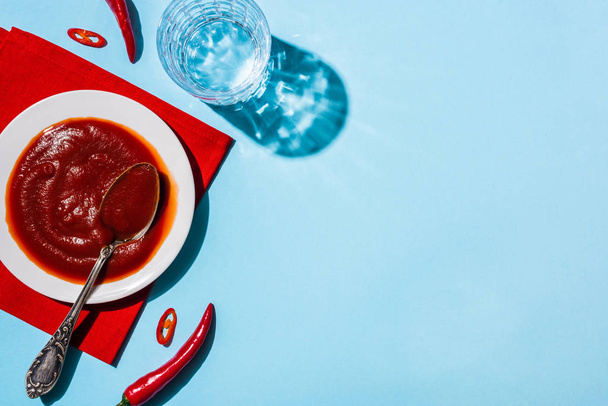 Delicious chili sauce in pate on red napkin beside glass of water and chili peppers on blue surface - Fotoğraf, Görsel