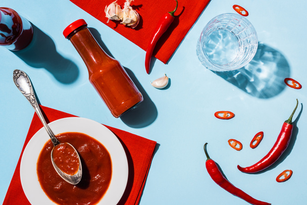 Top view of tasty tomato sauce with garlic and chili pepper beside glass of water on blue surface - 写真・画像