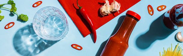 Вид сверху на кетчуп с чесноком, перцем чили и стаканом воды на голубом фоне, панорамный снимок
 - Фото, изображение