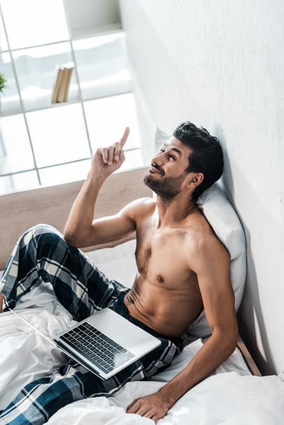 high angle view of handsome and smiling bi-racial man with laptop showing idea gesture in morning  - Foto, imagen