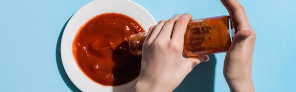 Cropped view of woman pouring tasty ketchup from bottle to plate on blue surface, panoramic shot - Φωτογραφία, εικόνα