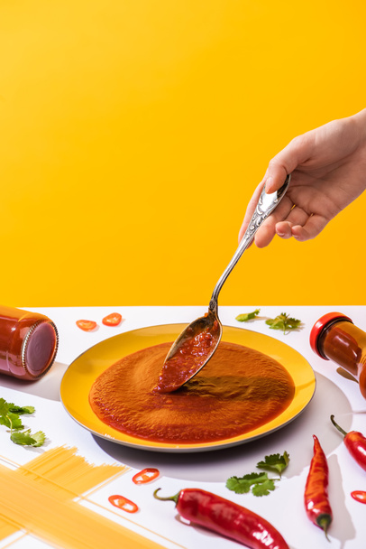 Cropped view of woman holding spoon with ketchup with spaghetti and chili peppers on white surface isolated on yellow - Photo, Image