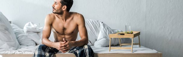 panoramic shot of handsome bi-racial man sitting on bed in morning  - 写真・画像
