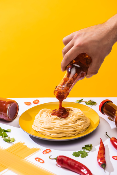 Cropped view of man pouring ketchup on spaghetti with chili peppers and greenery on white surface isolated on yellow - Foto, Imagen