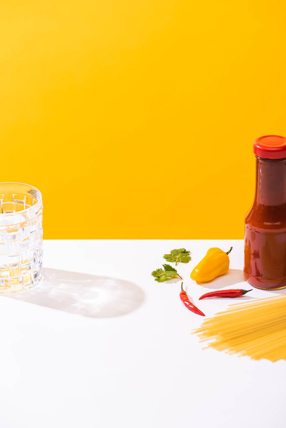 Bottle of ketchup beside peppers, raw spaghetti and glass of water on white surface on yellow background - Photo, Image