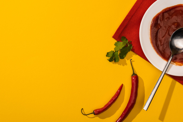 Top view of ketchup in plate with cilantro and chili peppers on yellow background - Photo, Image