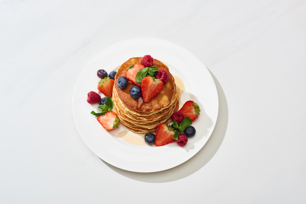 top view of delicious pancakes with honey, blueberries and strawberries on plate on marble white surface - Valokuva, kuva