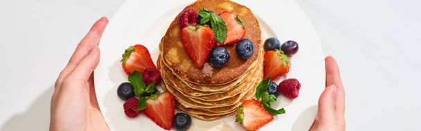cropped view of woman holding plate with delicious pancakes with honey, blueberries and strawberries, panoramic shot - Photo, Image