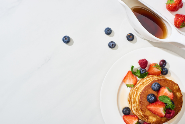 top view of delicious pancakes with maple syrup, blueberries and strawberries on plate on marble white surface - Valokuva, kuva