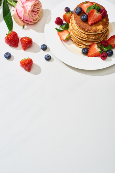 Blick von oben auf leckere Pfannkuchen mit Blaubeeren und Erdbeeren auf Teller in der Nähe von Rosenblüte auf weißem Marmor - Foto, Bild
