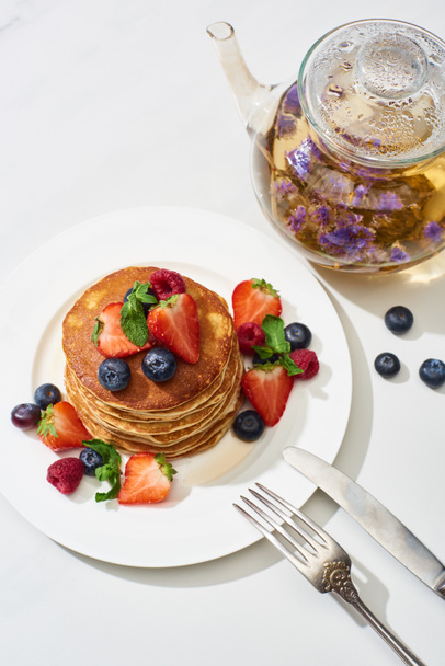 top view of delicious pancakes with maple syrup, blueberries and strawberries on plate near herbal tea in teapot, fork and knife on marble white surface - Photo, Image