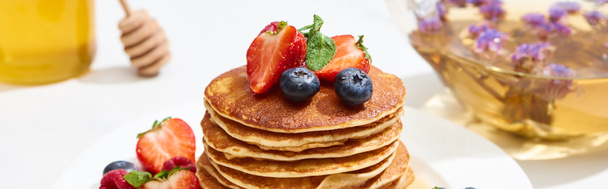 selektiver Fokus von leckeren Pfannkuchen mit Honig, Blaubeeren und Erdbeeren auf Teller auf weißer Oberfläche, Panoramaaufnahme - Foto, Bild