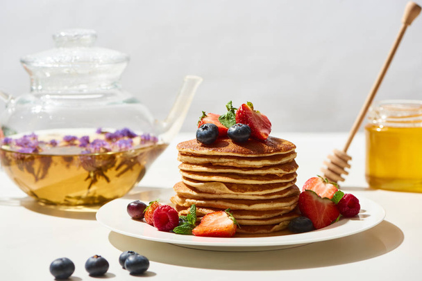 selective focus of delicious pancakes with honey, blueberries and strawberries on plate near herbal tea on white surface isolated on grey - Photo, Image