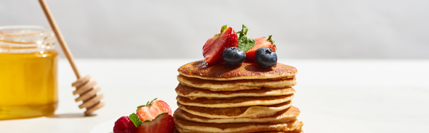 selektiver Fokus von leckeren Pfannkuchen mit Blaubeeren und Erdbeeren in der Nähe von Honig auf weißer Oberfläche isoliert auf grauer, panoramischer Aufnahme - Foto, Bild