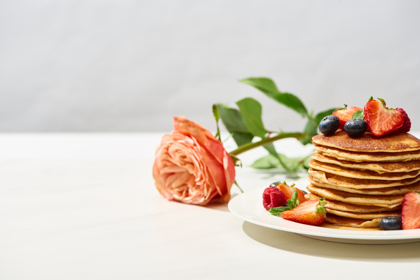 selective focus of delicious pancakes with blueberries and strawberries on plate near rose on white surface isolated on grey - Photo, Image