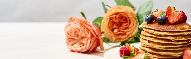 selectieve focus van heerlijke pannenkoeken met bosbessen en aardbeien op bord in de buurt van rozenbloemen op wit oppervlak geïsoleerd op grijze, panoramische opname - Foto, afbeelding