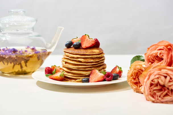 selective focus of delicious pancakes with blueberries and strawberries on plate near rose flowers and herbal tea on white surface isolated on grey - Fotoğraf, Görsel