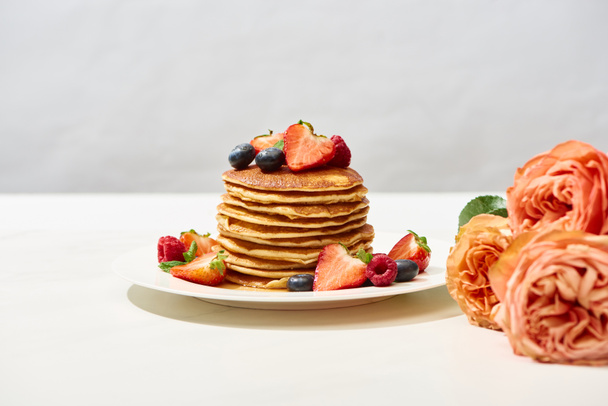 selective focus of delicious pancakes with blueberries and strawberries on plate near rose flowers on white surface isolated on grey - Photo, Image