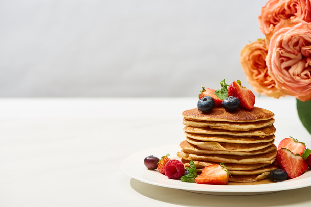 delicious pancakes with blueberries and strawberries on plate near rose flowers on white surface isolated on grey - Photo, Image
