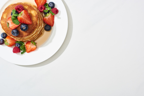 top view of delicious pancakes with honey, blueberries and strawberries on plate on marble white surface - Photo, Image