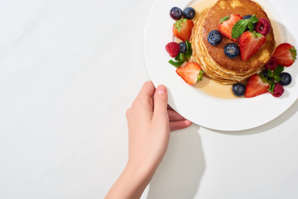 gedeeltelijk zicht op vrouw met bord met heerlijke pannenkoeken met honing, bosbessen en aardbeien op marmeren wit oppervlak - Foto, afbeelding