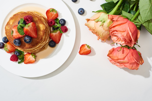 top view of delicious pancakes with honey, blueberries and strawberries on plate near roses on marble white surface - Photo, image